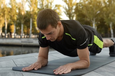 Sporty man doing plank exercise on mat near river