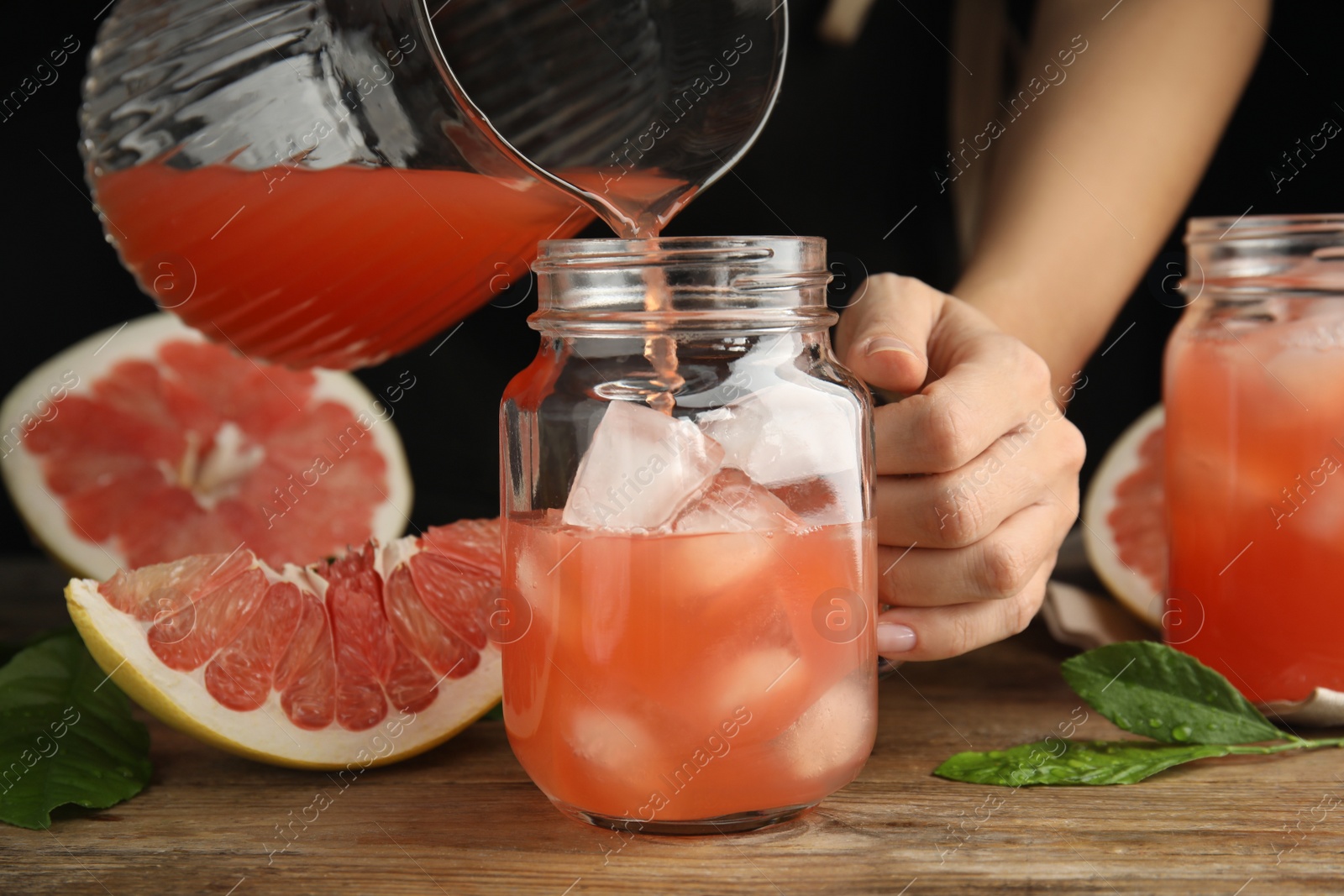Photo of Woman pouring pink pomelo juice into glass jar with ice at wooden table, closeup