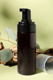 Bottle with face cleansing product, eucalyptus leaves and log on light green background, closeup