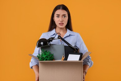 Unemployment problem. Confused woman with box of personal office belongings on orange background
