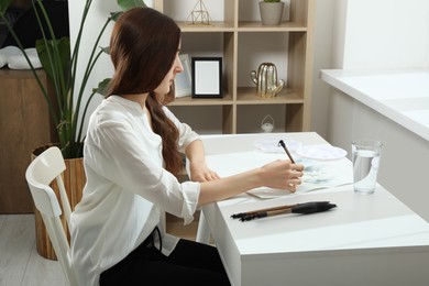 Photo of Woman painting flowers with watercolor at white table in room. Creative artwork