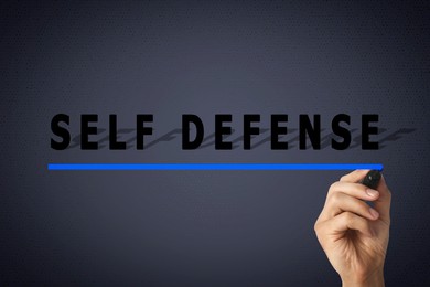 Man underlining words Self Defense with marker glass board against dark grey background, closeup