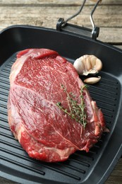 Photo of Grill pan with piece of raw beef meat, garlic and thyme on wooden table
