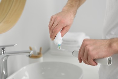 Man squeezing toothpaste from tube onto electric toothbrush above sink in bathroom, closeup