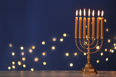 Golden menorah with burning candles on table against blue background and blurred festive lights, space for text