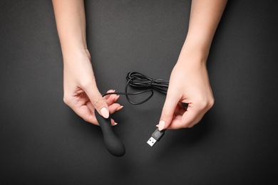 Photo of Young woman holding little vibrator with charging cable on black background, top view. Sex toy