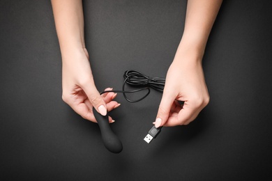 Young woman holding little vibrator with charging cable on black background, top view. Sex toy