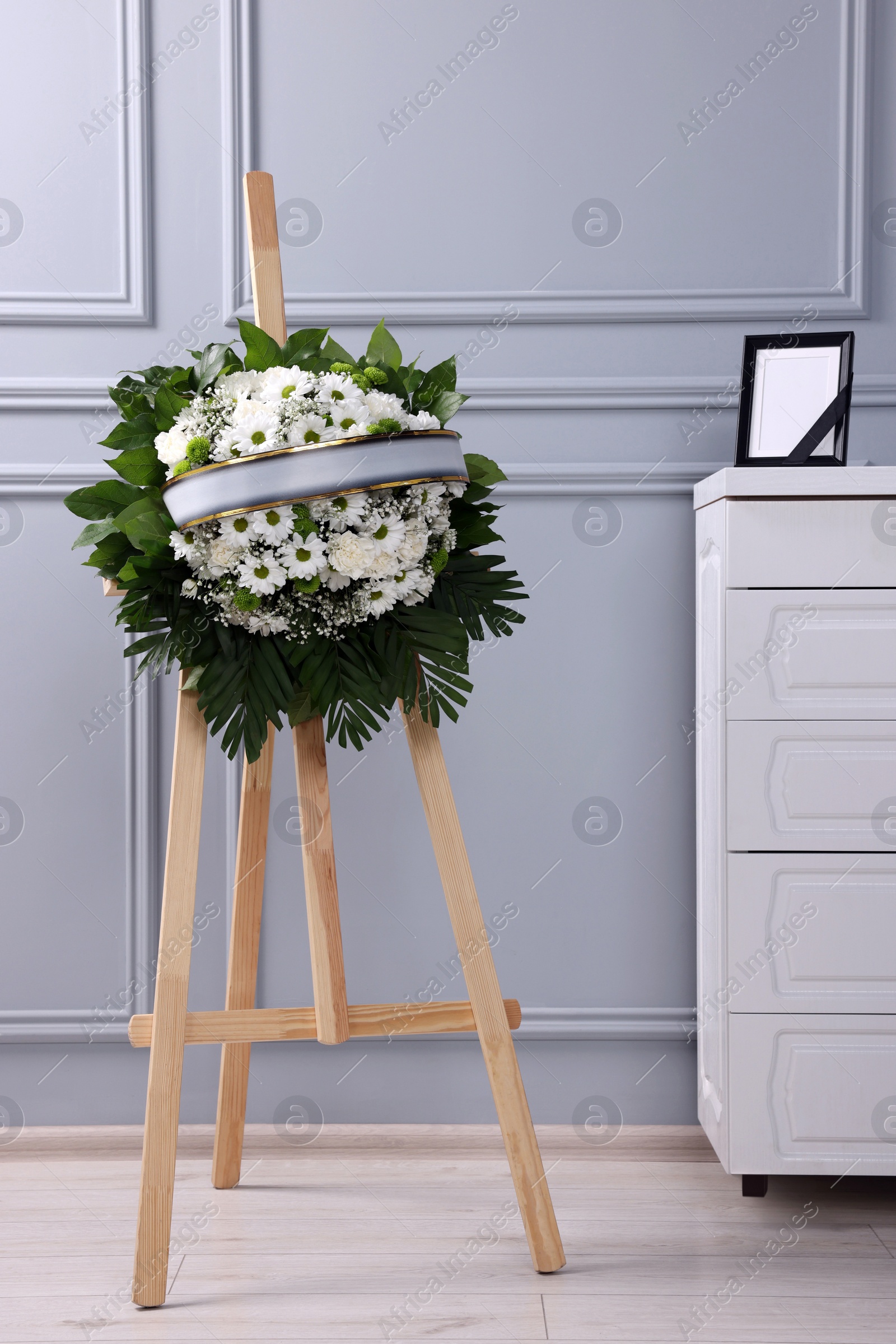 Photo of Funeral wreath of flowers on wooden stand and photo frame with black ribbon on white commode indoors