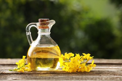 Photo of Rapeseed oil in glass jug and beautiful yellow flowers on wooden table outdoors