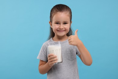 Photo of Cute girl with glass of fresh milk showing thumb up on light blue background