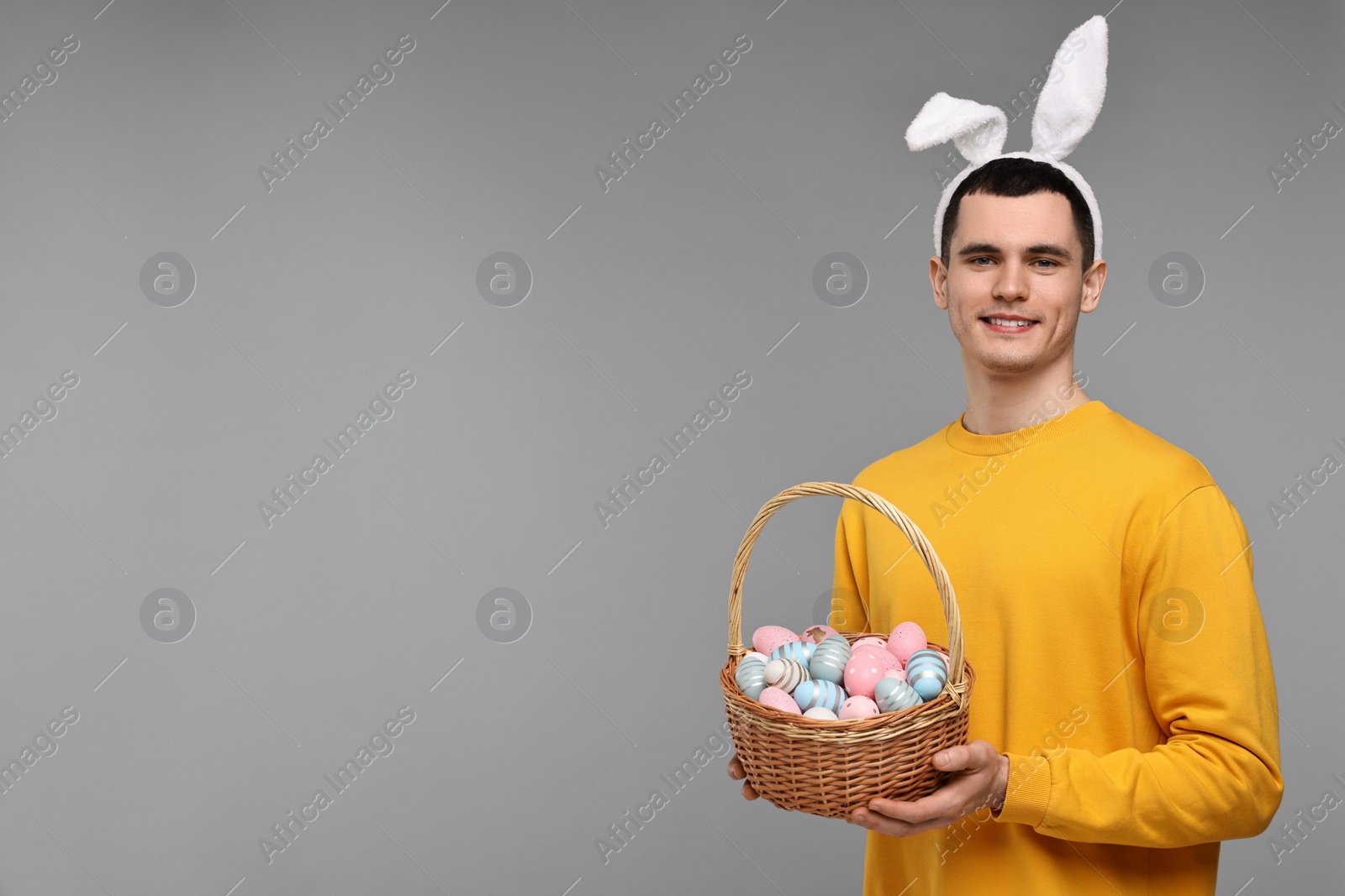 Photo of Easter celebration. Handsome young man with bunny ears holding basket of painted eggs on grey background. Space for text