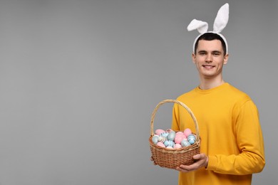 Photo of Easter celebration. Handsome young man with bunny ears holding basket of painted eggs on grey background. Space for text