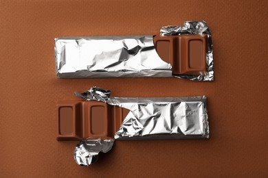 Photo of Delicious chocolate bars wrapped in foil on brown background, flat lay