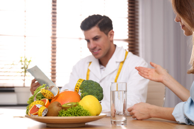 Nutritionist consulting patient at table in clinic, focus on plate with fruits, vegetables and measuring tape