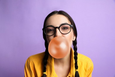 Photo of Fashionable young woman with braids blowing bubblegum on lilac background