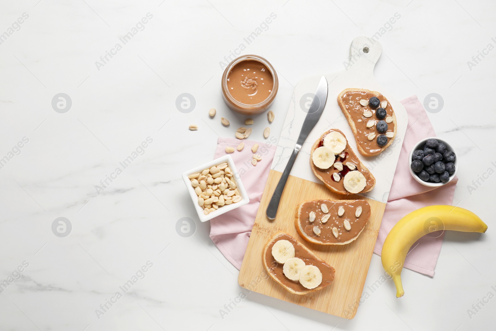 Photo of Toasts with tasty nut butter, banana slices, blueberries and peanuts on white marble table, flat lay. Space for text