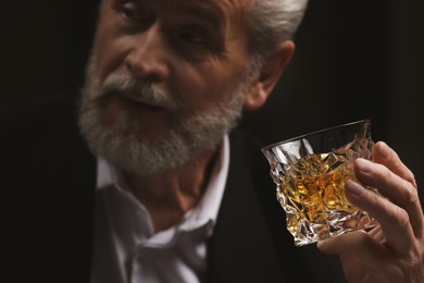 Man in suit holding glass of whiskey with ice cubes on black background, selective focus