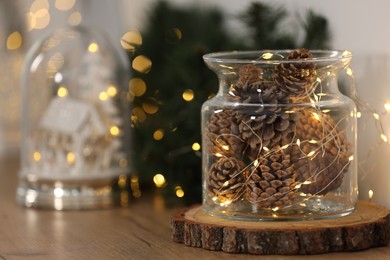 Photo of Christmas decoration. Cones and festive lights in jar on wooden table, closeup. Space for text