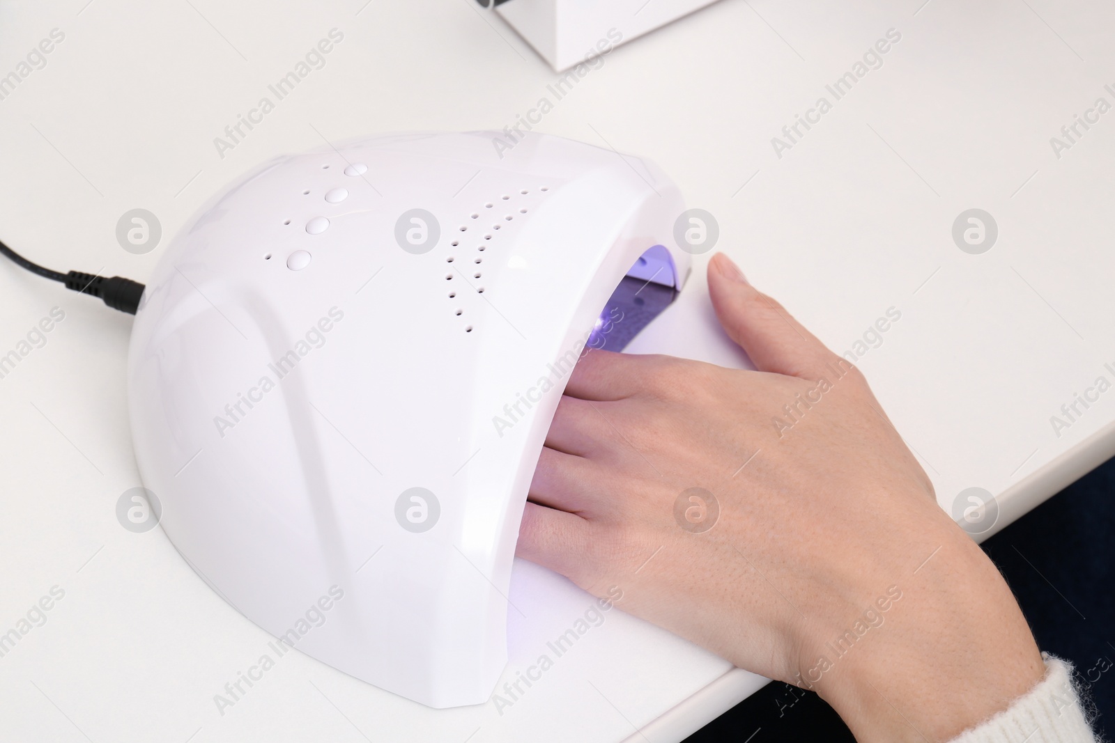 Photo of Woman using ultraviolet lamp to dry gel nail polish at white table, closeup