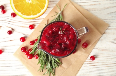 Tasty cranberry sauce in pitcher with rosemary on white wooden background, flat lay