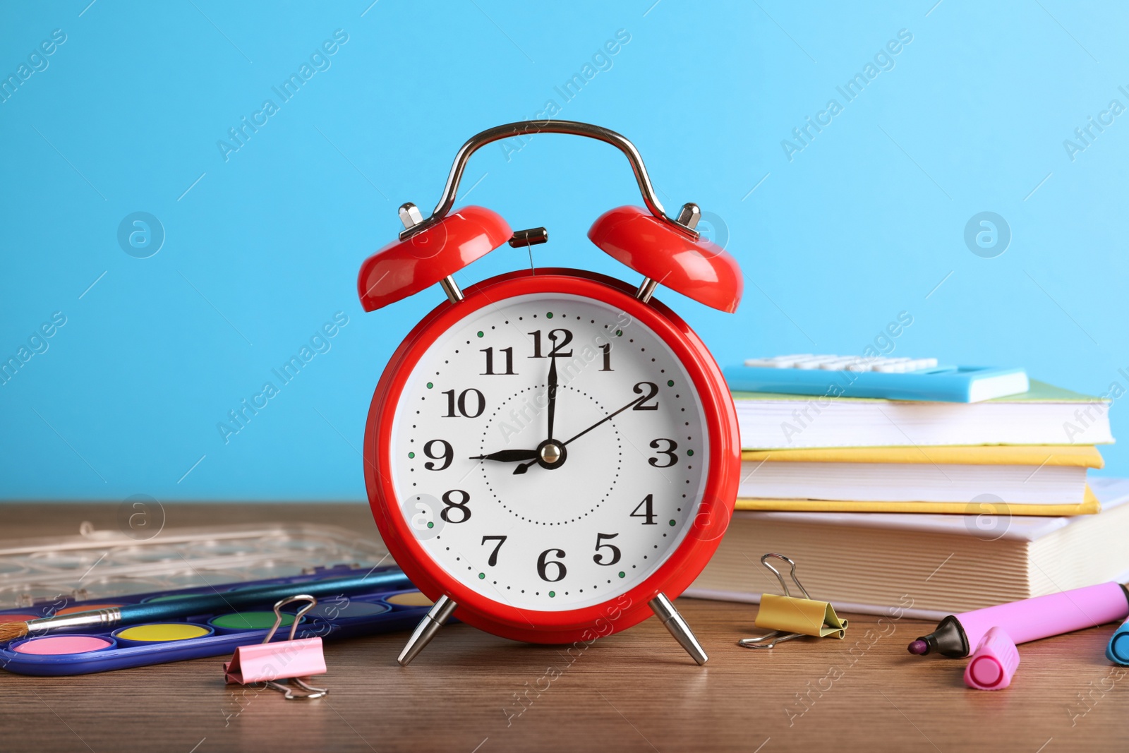 Photo of Red alarm clock and different stationery on wooden table against light blue background. School time