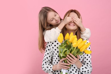 Photo of Daughter covering mother's eyes with her palms on pink background, space for text. Woman holding bouquet of yellow tulips