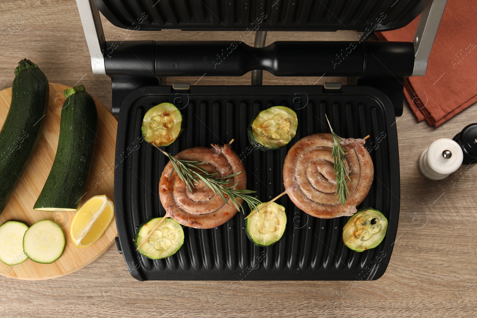 Photo of Electric grill with homemade sausages, zucchini and rosemary on wooden table, flat lay