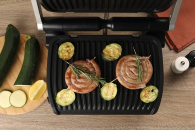 Photo of Electric grill with homemade sausages, zucchini and rosemary on wooden table, flat lay