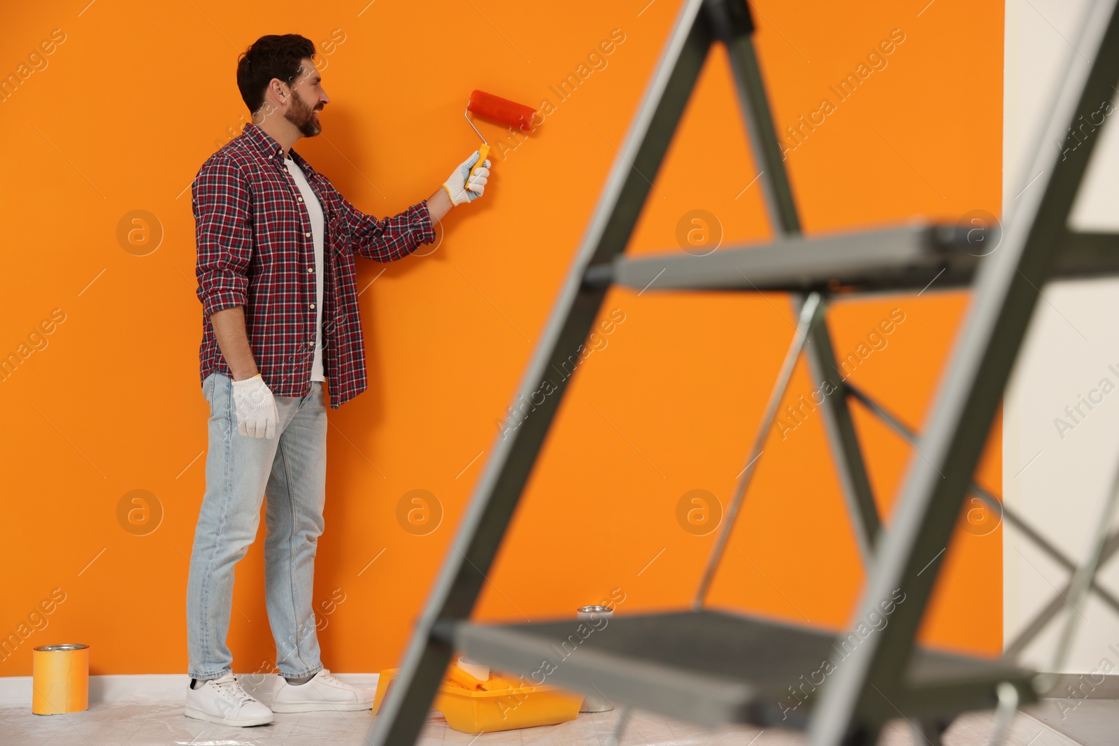 Photo of Designer painting orange wall with roller near folding ladder indoors