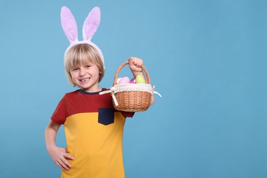Happy boy in bunny ears headband holding wicker basket with painted Easter eggs on turquoise background. Space for text