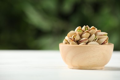 Tasty pistachios in bowl on white wooden table against blurred background, closeup. Space for text