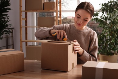 Young woman using utility knife to open parcel at wooden table indoors