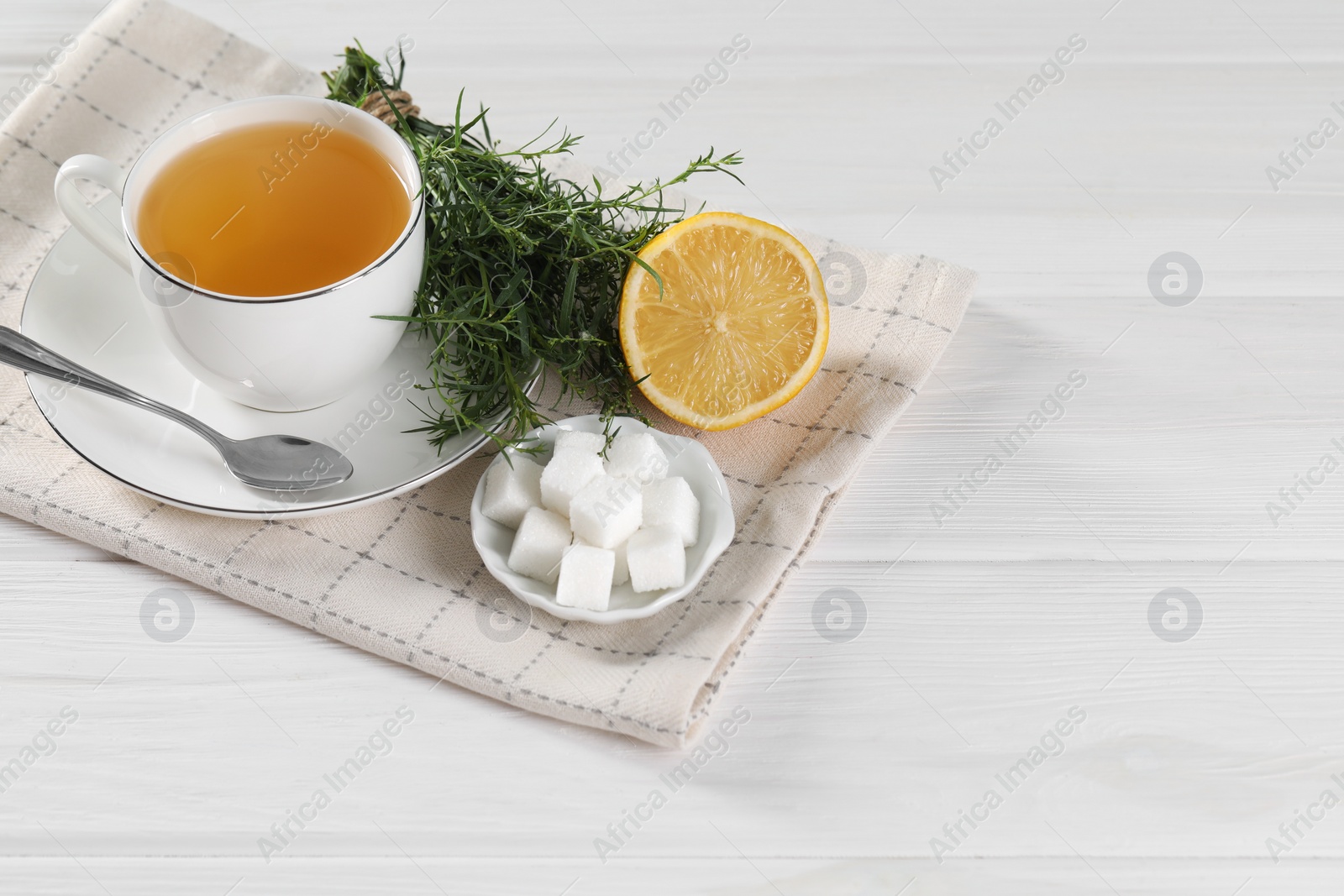 Photo of Aromatic herbal tea, fresh tarragon sprigs, sugar cubes and lemon on white wooden table, space for text