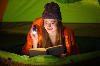 Photo of Young woman with flashlight reading book in tent