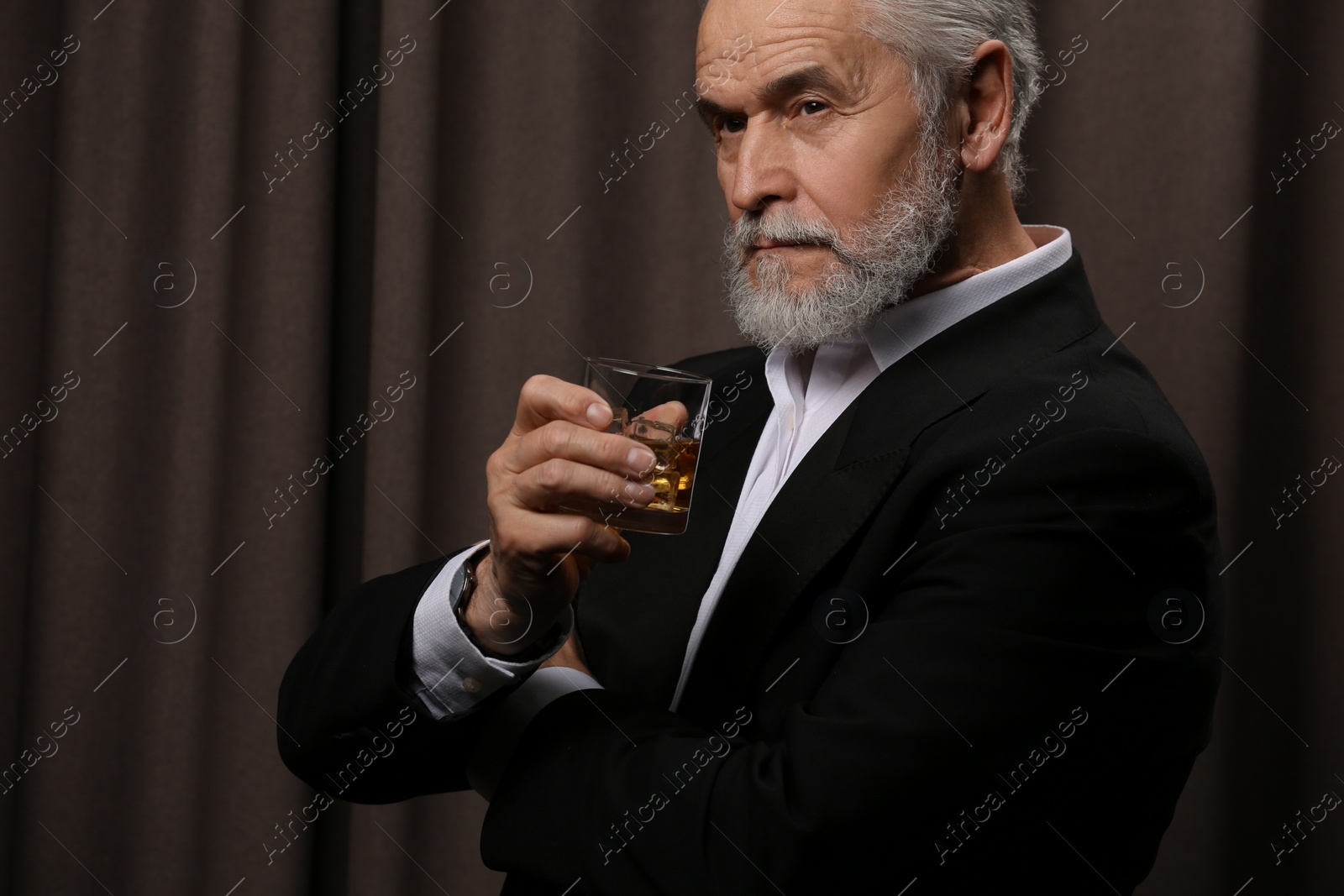 Photo of Senior man in suit holding glass of whiskey with ice cubes on brown background. Space for text