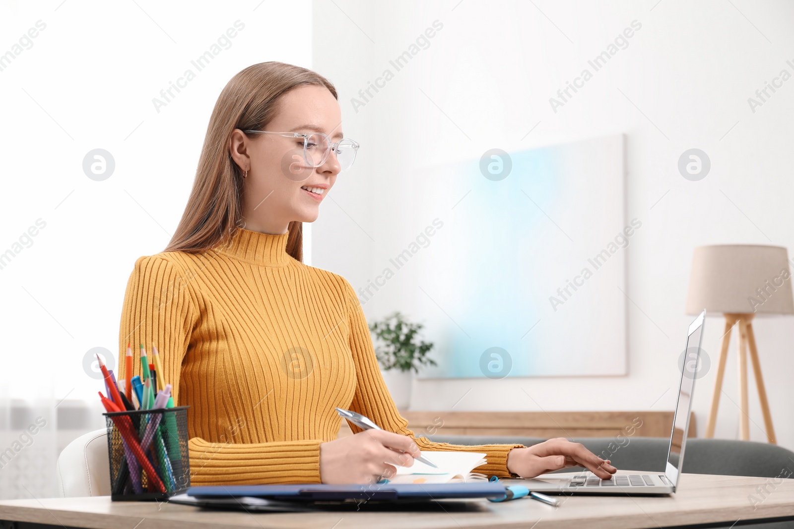 Photo of Woman taking notes while using laptop at wooden table in office