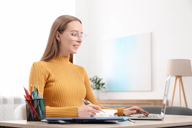 Photo of Woman taking notes while using laptop at wooden table in office