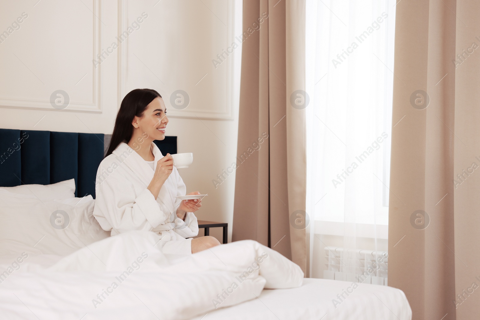 Photo of Happy young woman wearing bathrobe drinking coffee on bed in hotel room