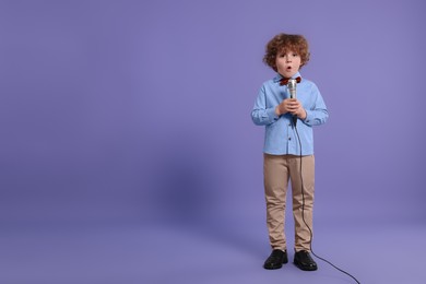 Photo of Cute little boy with microphone singing on violet background, space for text
