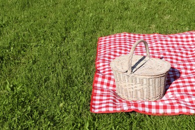 Photo of Picnic basket with checkered tablecloth on green grass outdoors, space for text