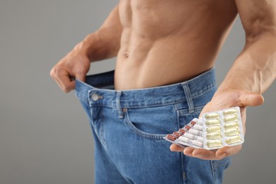 Athletic man with pills wearing big jeans on grey background, closeup. Weight loss