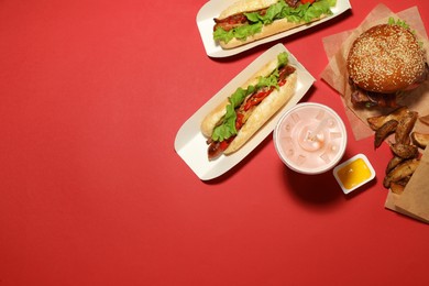 Tasty burger, hot dogs, potato wedges, sauce and refreshing drink on red background, flat lay with space for text. Fast food