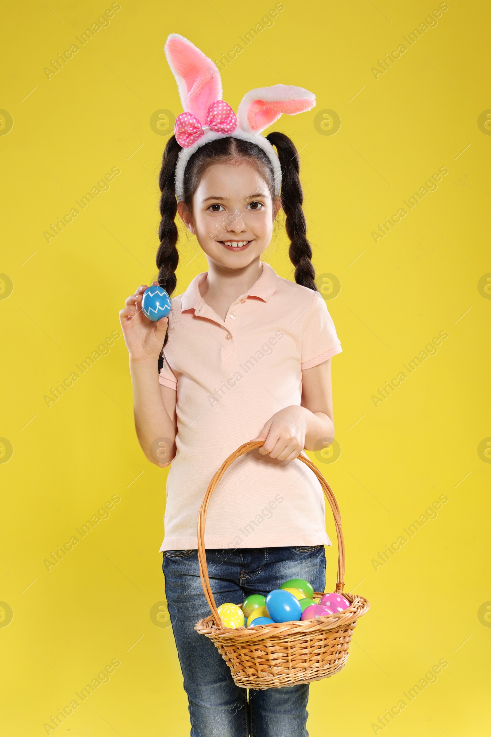 Photo of Little girl in bunny ears headband holding basket with Easter eggs on color background