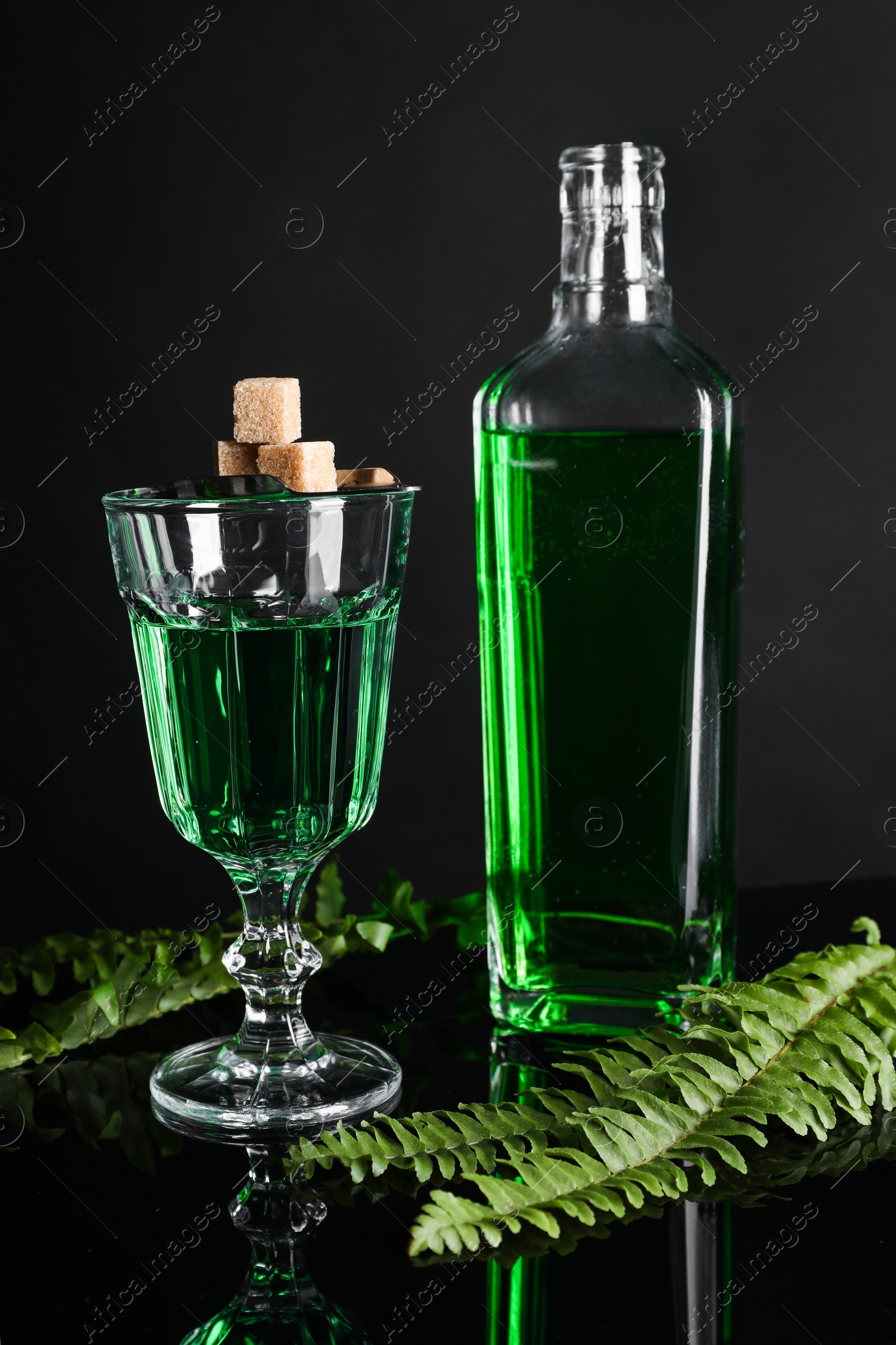 Photo of Absinthe, brown sugar, spoon and green leaves on mirror table. Alcoholic drink