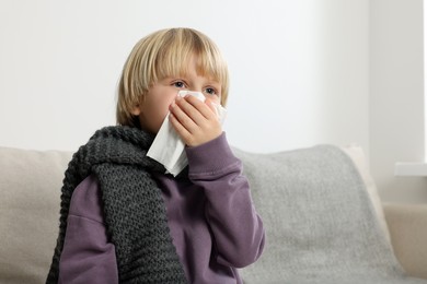 Photo of Boy blowing nose in tissue on sofa in room. Cold symptoms