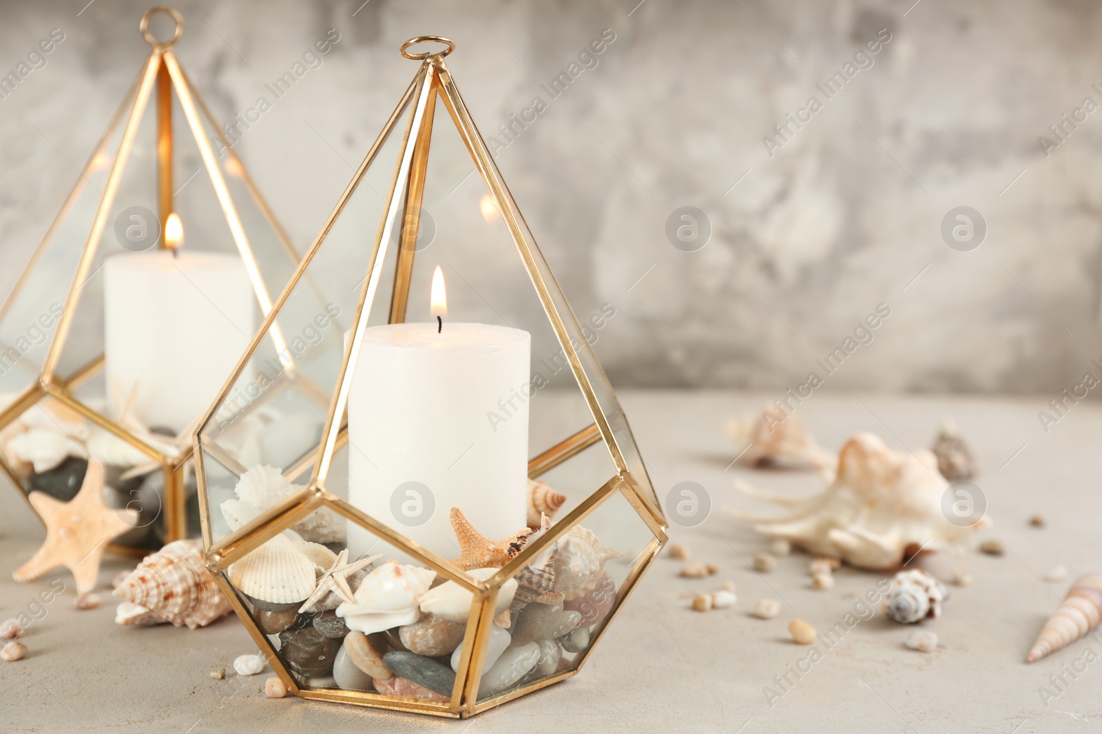 Photo of Stylish glass holders with burning candles, seashells and pebbles on light stone table