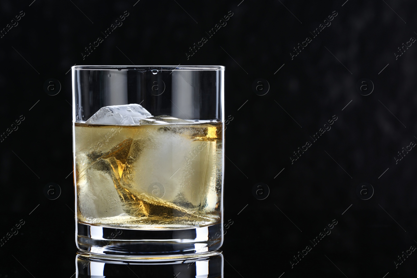 Photo of Tasty whiskey and ice cubes in glass on mirror table against black background, closeup. Space for text