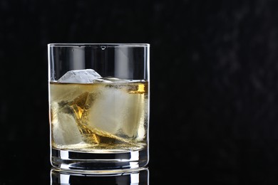 Photo of Tasty whiskey and ice cubes in glass on mirror table against black background, closeup. Space for text