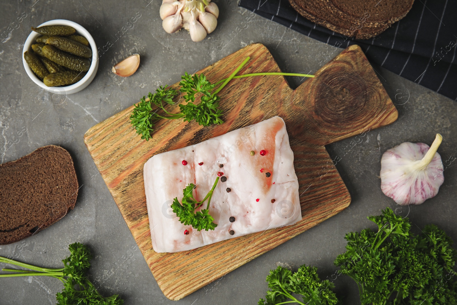 Photo of Board with pork fatback, garlic, parsley, bread and bowl of pickles on grey stone background, flat lay