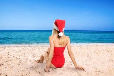 Young woman wearing Santa Claus hat on beach near sea, back view. Christmas vacation